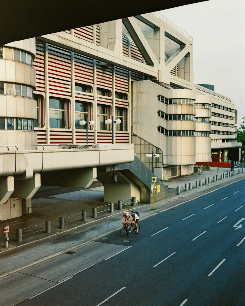 two cyclists on a street alongside a big, futuristic looking building