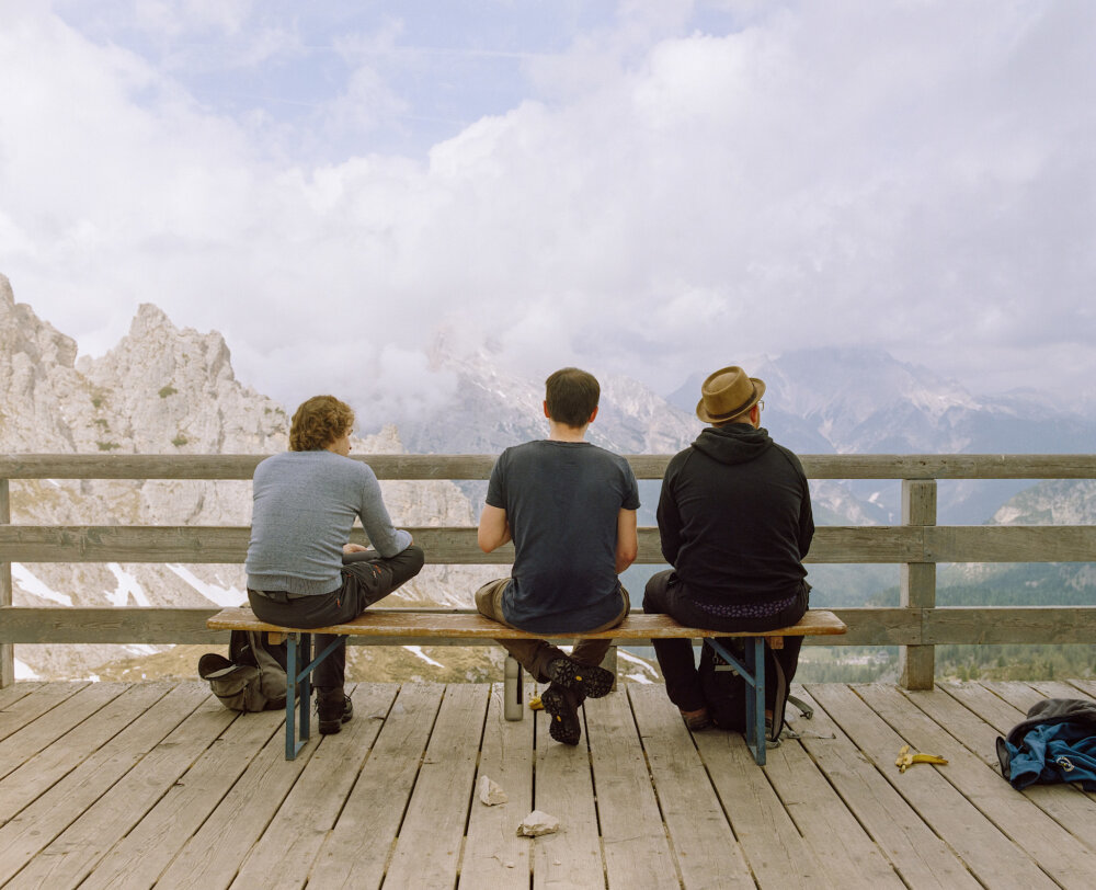 three men sitting on a wooden deck looking at a mountain panorama